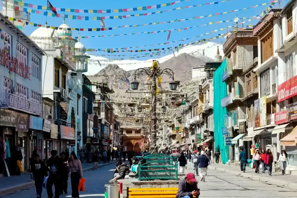 A vibrant view of the pedestrian-only Leh Main Market, bustling with visitors walking past traditional shops, with colorful prayer flags strung overhead and snow-capped mountains in the bright blue distance.