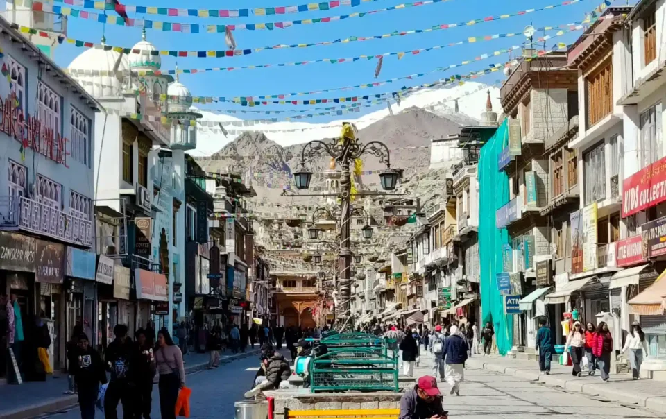 A vibrant view of the pedestrian-only Leh Main Market, bustling with visitors walking past traditional shops, with colorful prayer flags strung overhead and snow-capped mountains in the bright blue distance.