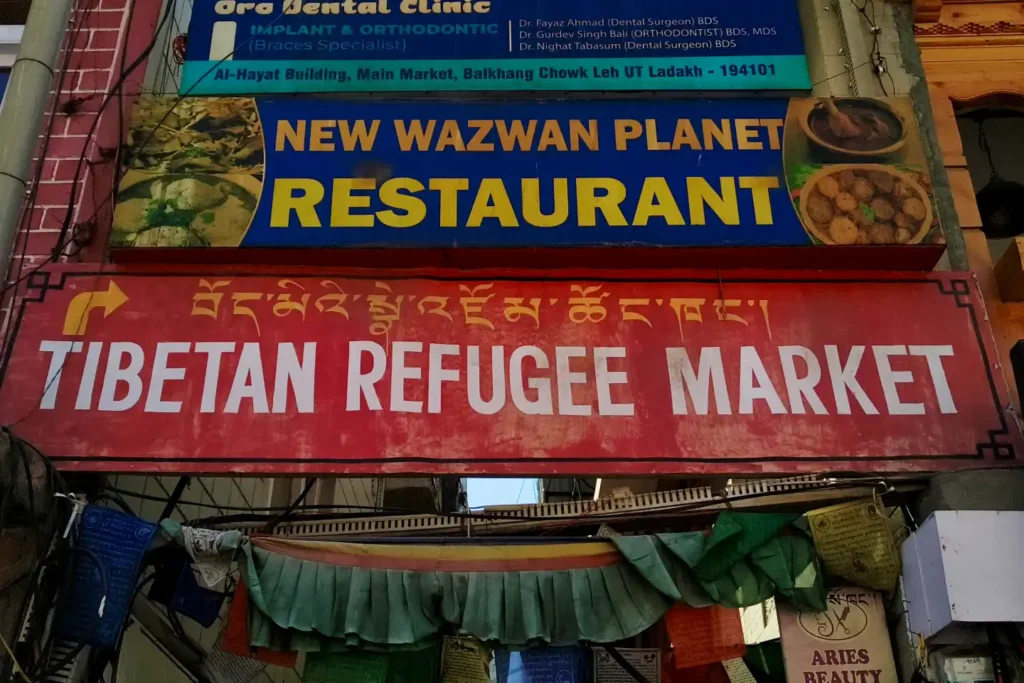 A prominent red and white signboard marking the entrance to the Tibetan Refugee Market in Leh, hanging on a brick building beneath a restaurant sign and above traditional draped fabrics.